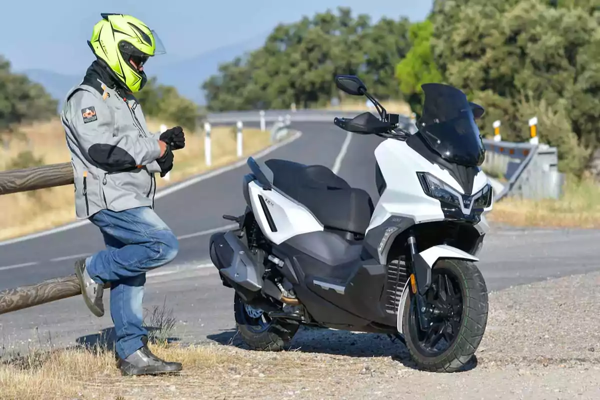 Persona con casco y chaqueta de motociclista apoyada en una cerca junto a una moto tipo scooter blanca en una carretera curva rodeada de vegetación Persona con casco y chaqueta de motociclista apoyada en una cerca junto a una moto tipo scooter blanca en una carretera curva rodeada de vegetación