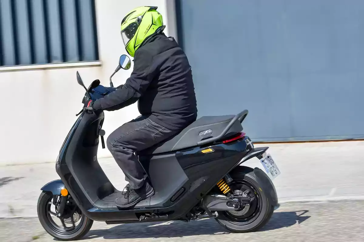 Persona con casco amarillo fosforescente y chaqueta negra conduciendo una moto tipo scooter negra en una calle urbana Persona con casco amarillo fosforescente y chaqueta negra conduciendo una moto tipo scooter negra en una calle urbana