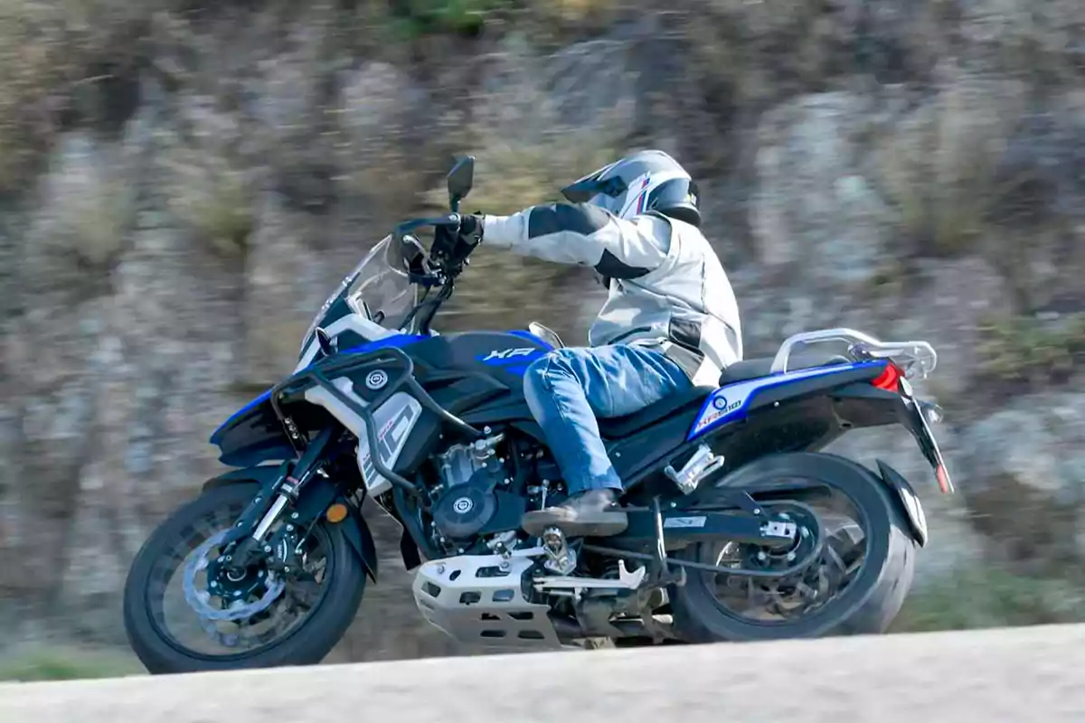 Persona conduint una motocicleta blava per una carretera amb fons de vegetació i roques Persona conduint una motocicleta blava per una carretera amb fons de vegetació i roques