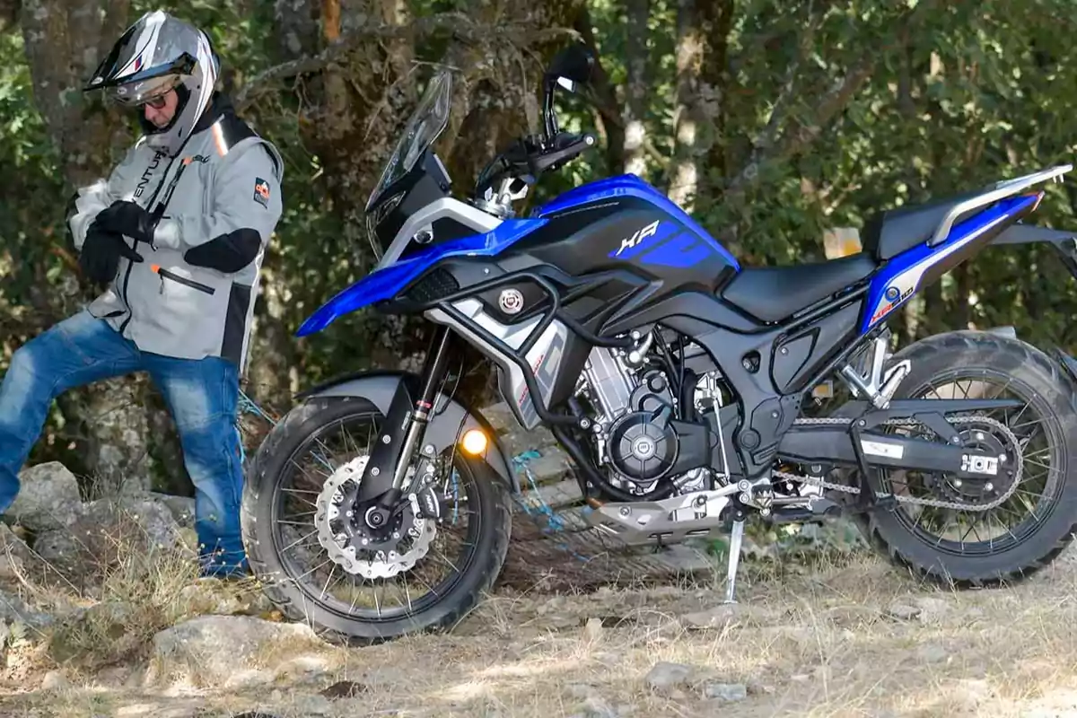 Motocicleta blava estacionada en un entorn natural amb una persona amb casc i jaqueta de protecció dreta al seu costat Motocicleta blava estacionada en un entorn natural amb una persona amb casc i jaqueta de protecció dreta al seu costat