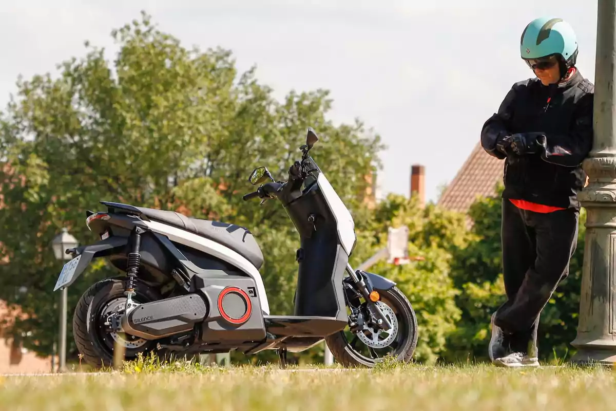 Persona con casco y chaqueta de motociclista de pie junto a una moto eléctrica en un entorno al aire libre con árboles al fondo Persona con casco y chaqueta de motociclista de pie junto a una moto eléctrica en un entorno al aire libre con árboles al fondo