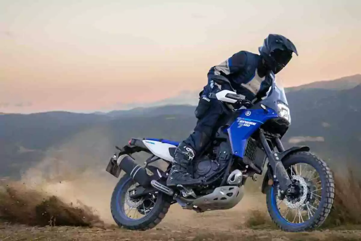 Persona conduciendo una motocicleta azul Yamaha en un camino de tierra con montañas al fondo al atardecer Persona conduciendo una motocicleta azul Yamaha en un camino de tierra con montañas al fondo al atardecer