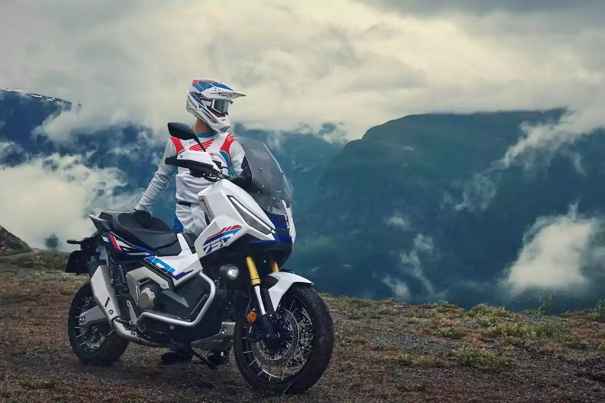 Persona con equipo de motocross junto a una motocicleta blanca en un paisaje montañoso con nubes bajas y vegetación alpina. Persona con equipo de motocross junto a una motocicleta blanca en un paisaje montañoso con nubes bajas y vegetación alpina.
