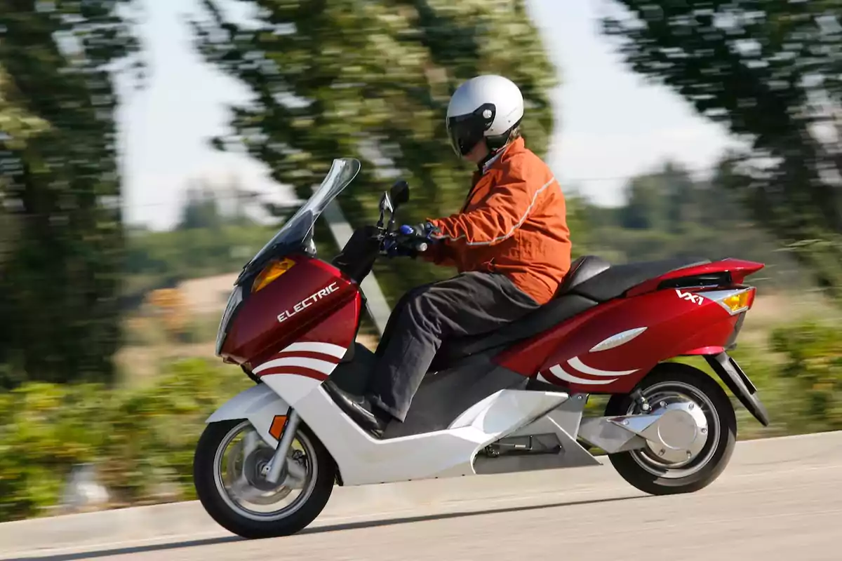 Persona conduciendo una moto eléctrica roja con casco blanco y chaqueta naranja en una carretera rodeada de árboles Persona conduciendo una moto eléctrica roja con casco blanco y chaqueta naranja en una carretera rodeada de árboles