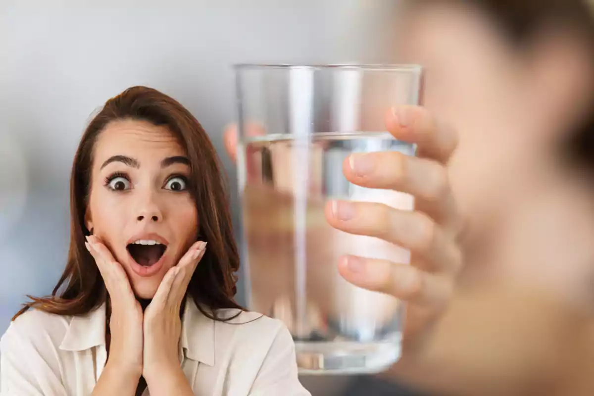 Mujer con expresión de sorpresa junto a un vaso de agua sostenido por una mano Mujer con expresión de sorpresa junto a un vaso de agua sostenido por una mano