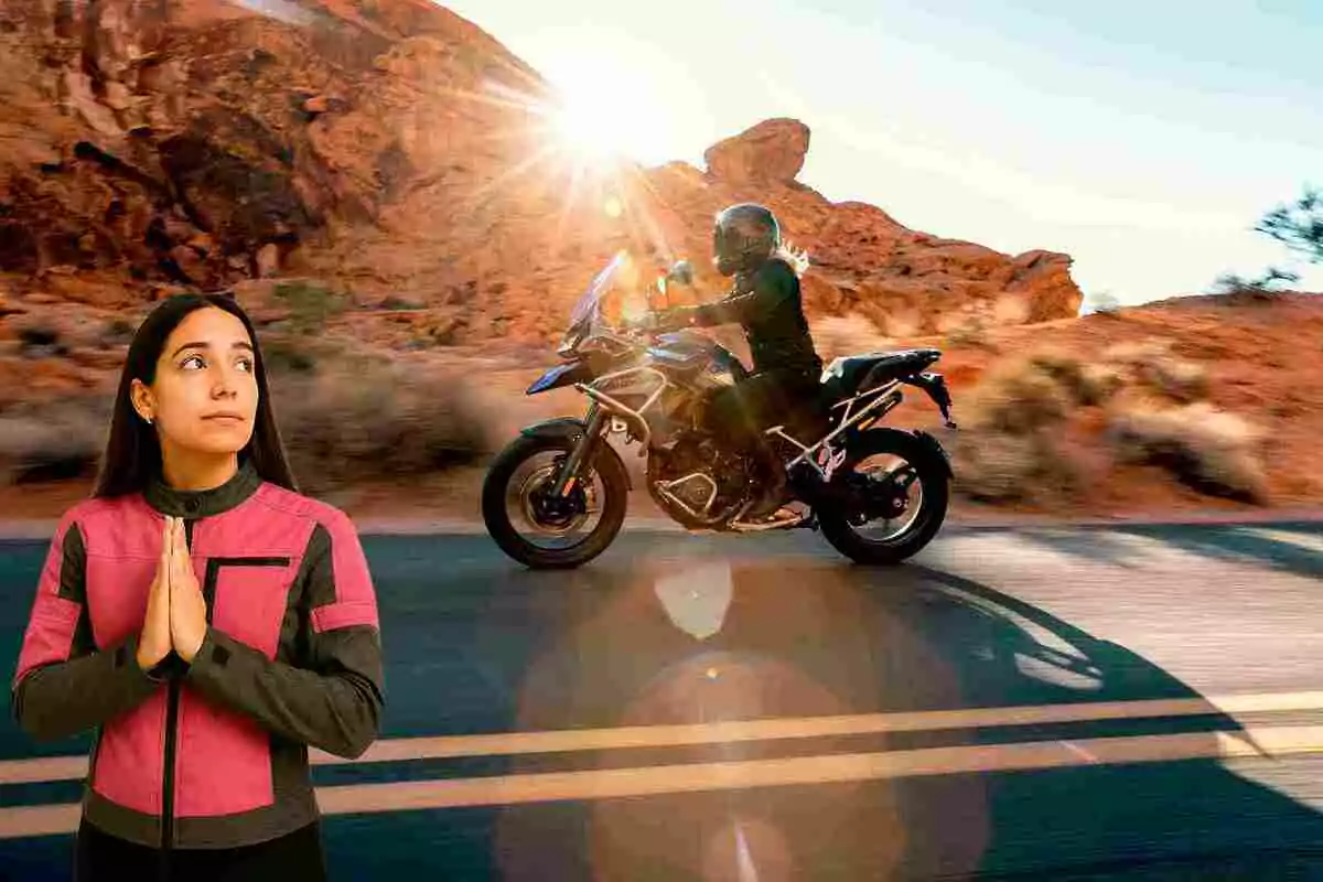 Woman in a pink and black jacket looking up with her hands together as a motorcycle passes by on a road in a desert landscape at sunset