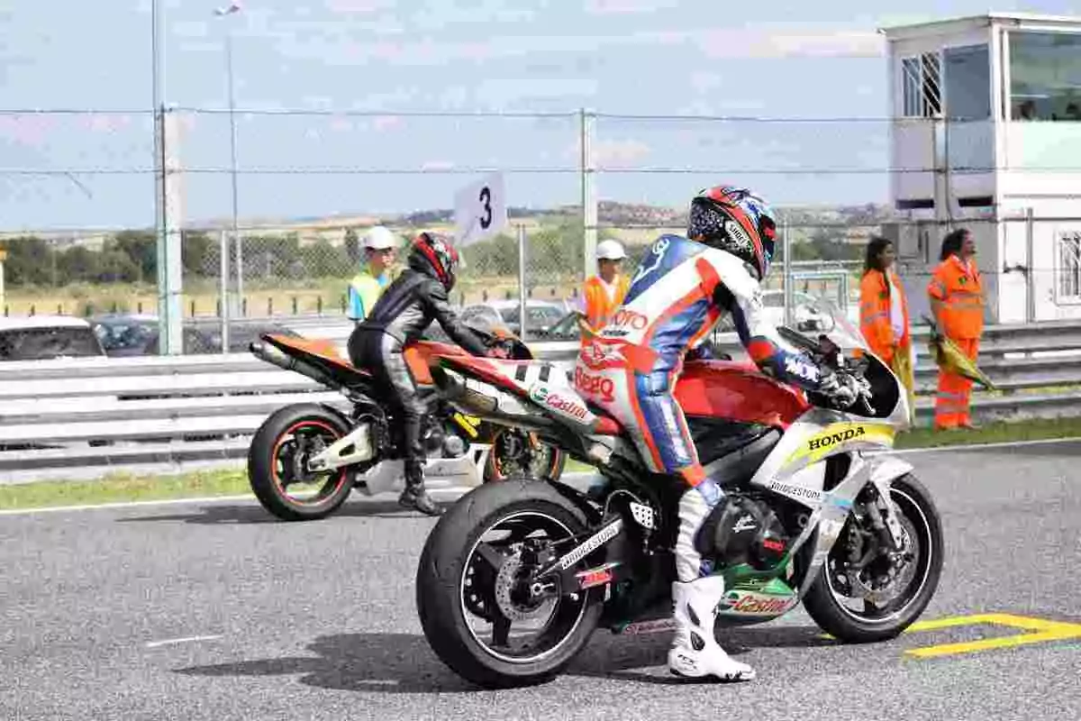 Two motorcyclists in racing suits are on the starting line of a track while several people in orange vests and uniforms watch from the background. Two motorcyclists in racing suits are on the starting line of a track while several people in orange vests and uniforms watch from the background.