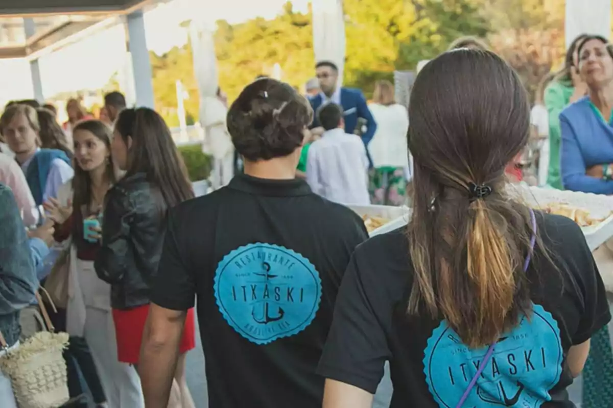 Dos camareros con camisetas negras que llevan el logo de un restaurante caminan entre un grupo de personas en un evento al aire libre Dos camareros con camisetas negras que llevan el logo de un restaurante caminan entre un grupo de personas en un evento al aire libre