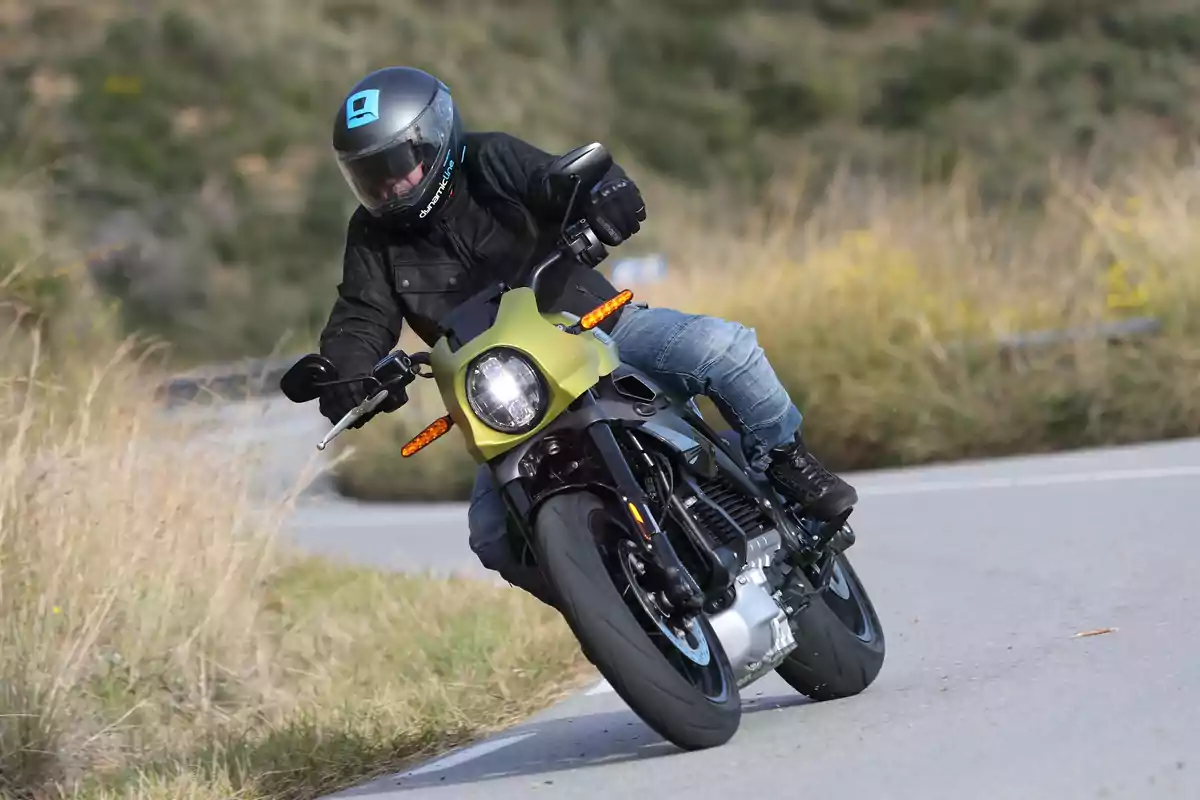 Persona con casco y chaqueta negra conduciendo una motocicleta en una carretera curva rodeada de vegetación Persona con casco y chaqueta negra conduciendo una motocicleta en una carretera curva rodeada de vegetación