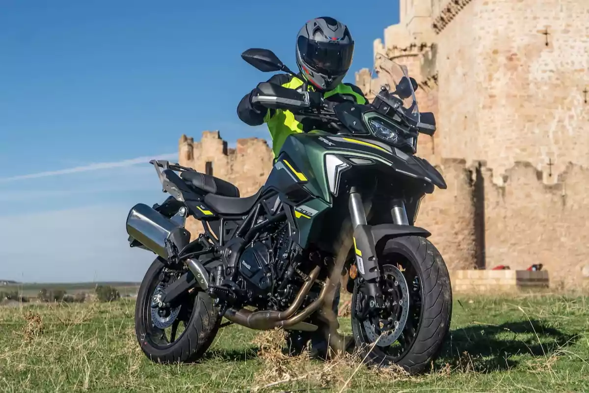 Motorista amb casc i jaqueta fosforescent posant damunt d’una motocicleta esportiva negra i verda davant d’una antiga construcció de pedra en un dia assolellat