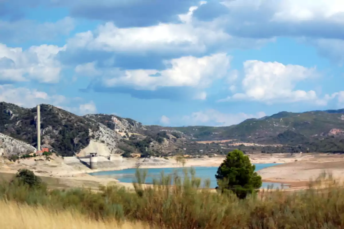 Paisaje de un embalse con poca agua rodeado de montañas y vegetación bajo un cielo parcialmente nublado Paisaje de un embalse con poca agua rodeado de montañas y vegetación bajo un cielo parcialmente nublado