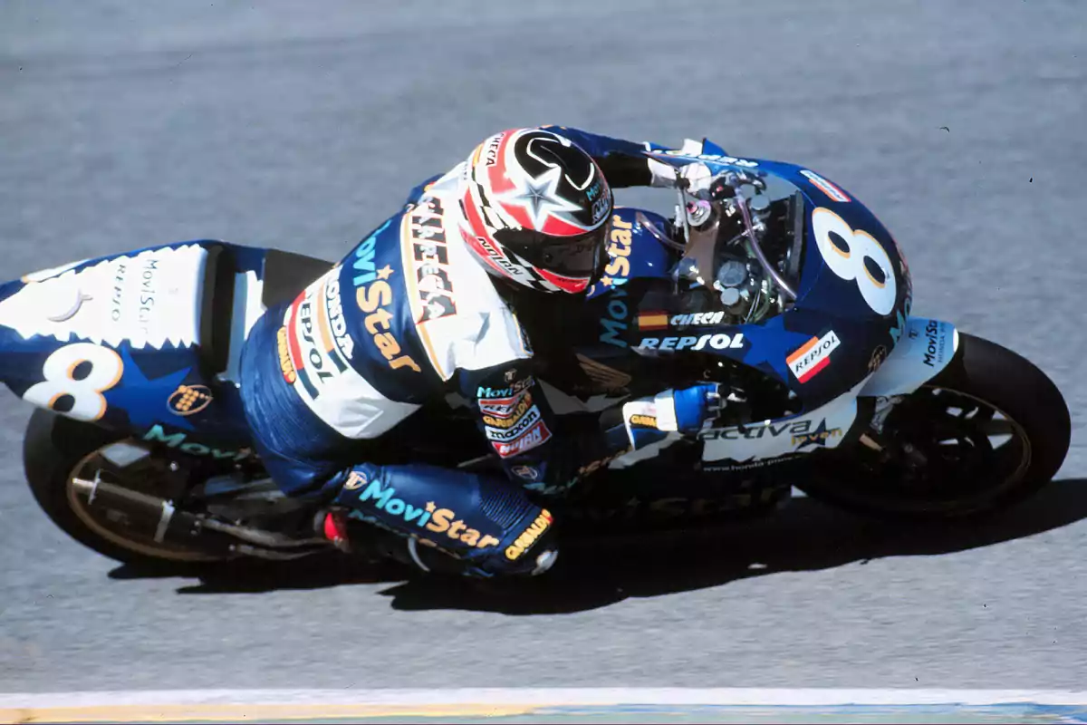 Carlos Checa tomando una curva en una pista de carreras con una moto azul decorada con el número 8 y patrocinadores visibles en el traje y la motocicleta Carlos Checa tomando una curva en una pista de carreras con una moto azul decorada con el número 8 y patrocinadores visibles en el traje y la motocicleta