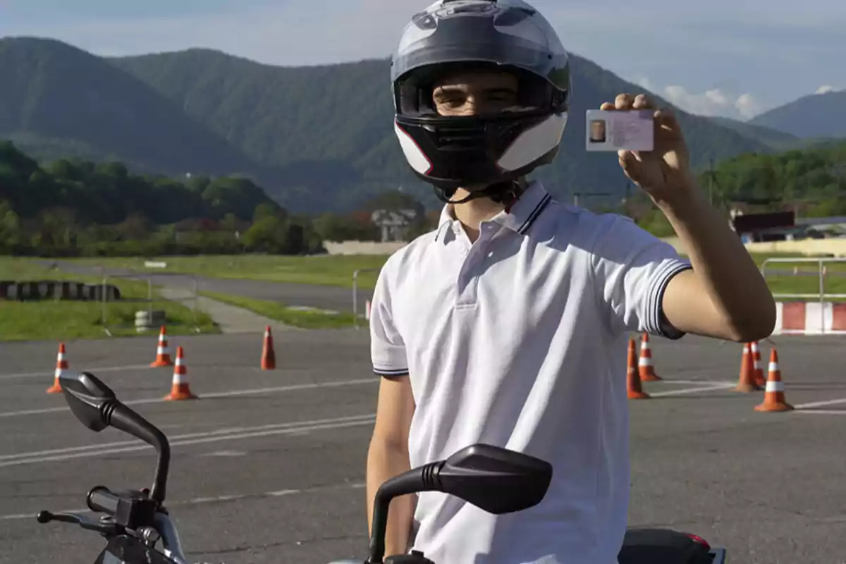 Joven con casco de motocicleta mostrando su licencia mientras está junto a una moto en una pista al aire libre con conos naranjas y montañas al fondo Joven con casco de motocicleta mostrando su licencia mientras está junto a una moto en una pista al aire libre con conos naranjas y montañas al fondo