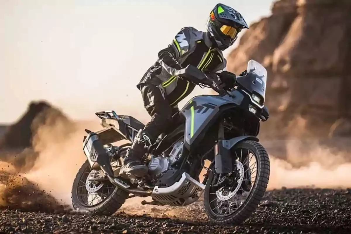 Persona conduint una motocicleta d’aventura en un terreny arenós amb equip de protecció i fons de roques i pols. Persona conduint una motocicleta d’aventura en un terreny arenós amb equip de protecció i fons de roques i pols.