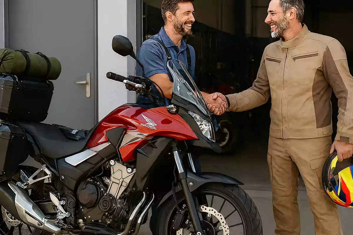 Dos hombres sonríen y se dan la mano junto a una motocicleta roja equipada para viajar, uno de ellos sostiene un casco multicolor