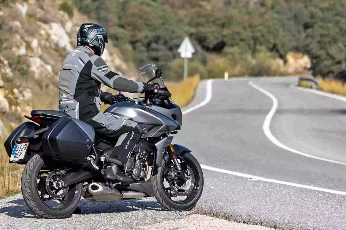 Person wearing a helmet and motorcycle suit sitting on a gray motorcycle at the edge of a curved road in a natural setting Person wearing a helmet and motorcycle suit sitting on a gray motorcycle at the edge of a curved road in a natural setting