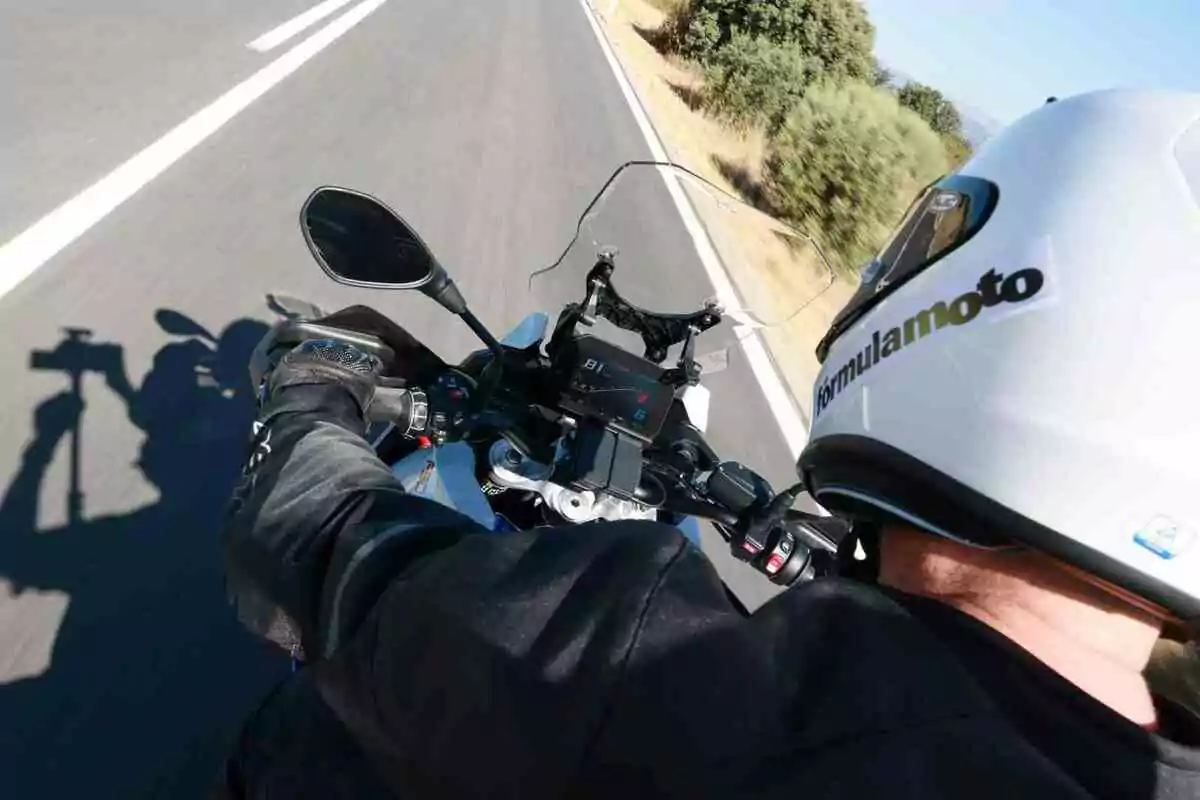Person riding a motorcycle on the road with a white helmet and black jacket, seen from behind and slightly above, showing the instrument panel and the landscape alongside the road Person riding a motorcycle on the road with a white helmet and black jacket, seen from behind and slightly above, showing the instrument panel and the landscape alongside the road