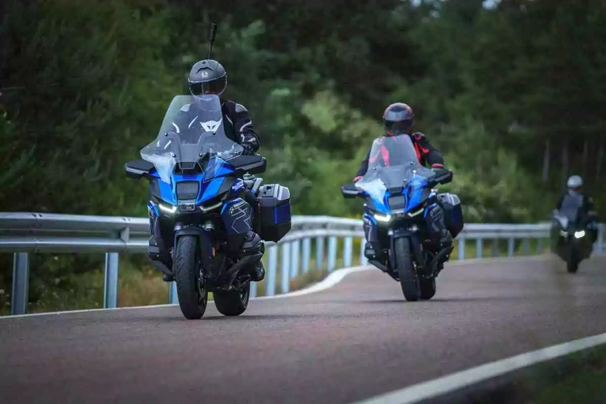 Two motorcyclists wearing protective gear ride blue sport bikes on a road surrounded by trees and metal guardrails. Two motorcyclists wearing protective gear ride blue sport bikes on a road surrounded by trees and metal guardrails.