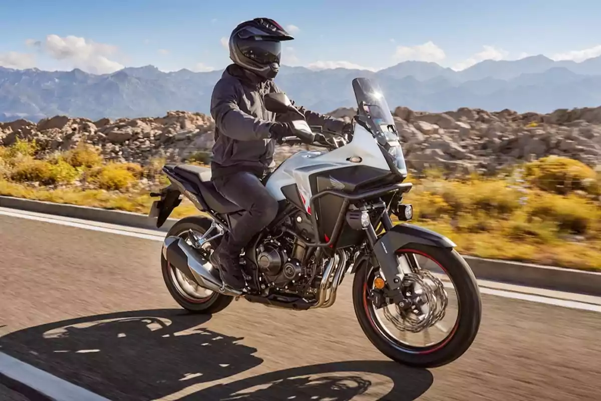 Persona conduint una motocicleta per una carretera envoltada de muntanyes i vegetació sota un cel serè Persona conduint una motocicleta per una carretera envoltada de muntanyes i vegetació sota un cel serè
