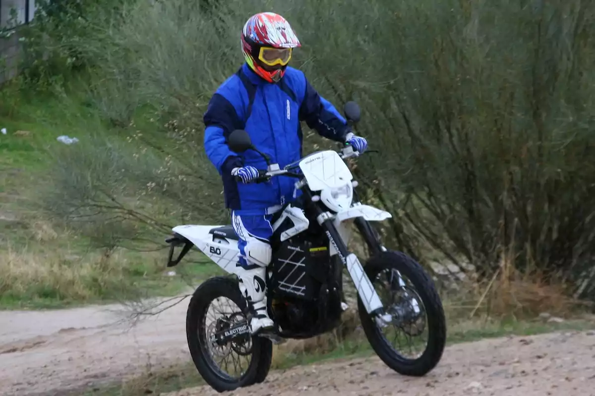 Persona con casco y ropa de protección azul conduciendo una motocicleta blanca por un camino de tierra rodeado de vegetación Persona con casco y ropa de protección azul conduciendo una motocicleta blanca por un camino de tierra rodeado de vegetación