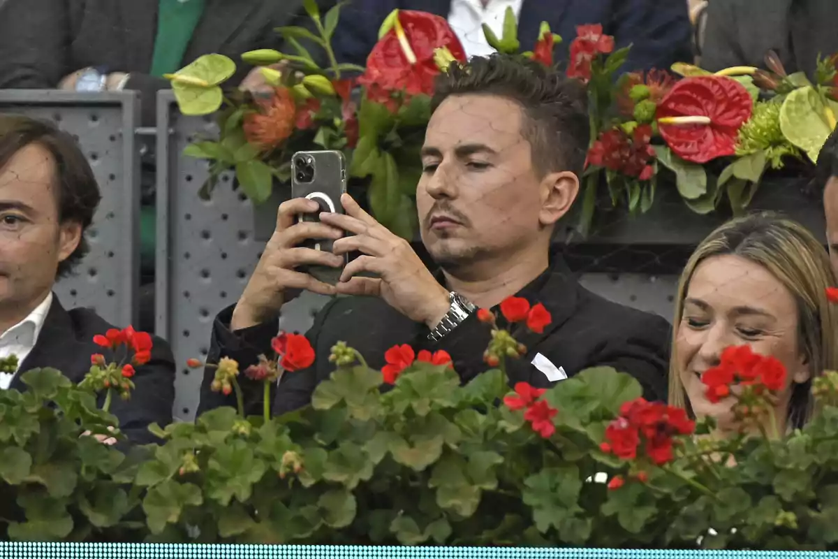 Jorge Lorenzo con traje negro tomando una foto con su celular entre flores rojas en un evento Jorge Lorenzo con traje negro tomando una foto con su celular entre flores rojas en un evento