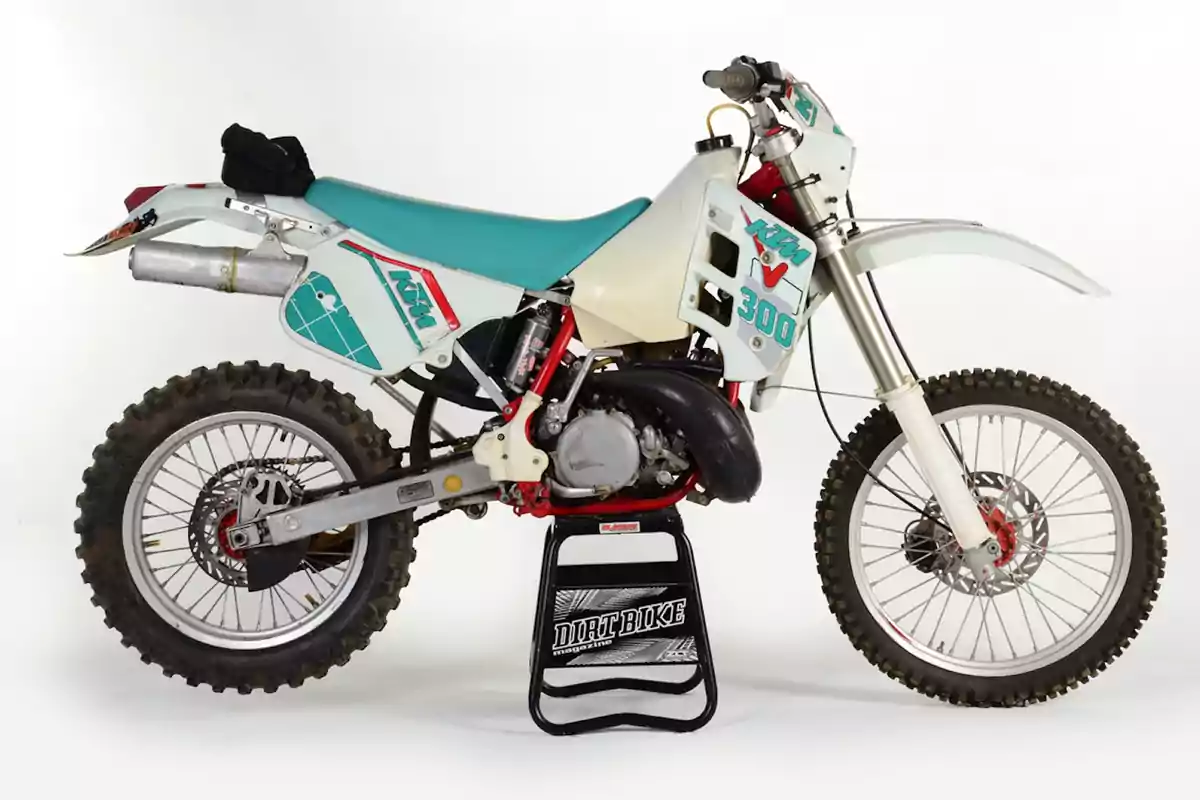 Motocicleta de motocross KTM 300 antigua con asiento turquesa y detalles en blanco y rojo sobre un soporte negro Motocicleta de motocross KTM 300 antigua con asiento turquesa y detalles en blanco y rojo sobre un soporte negro