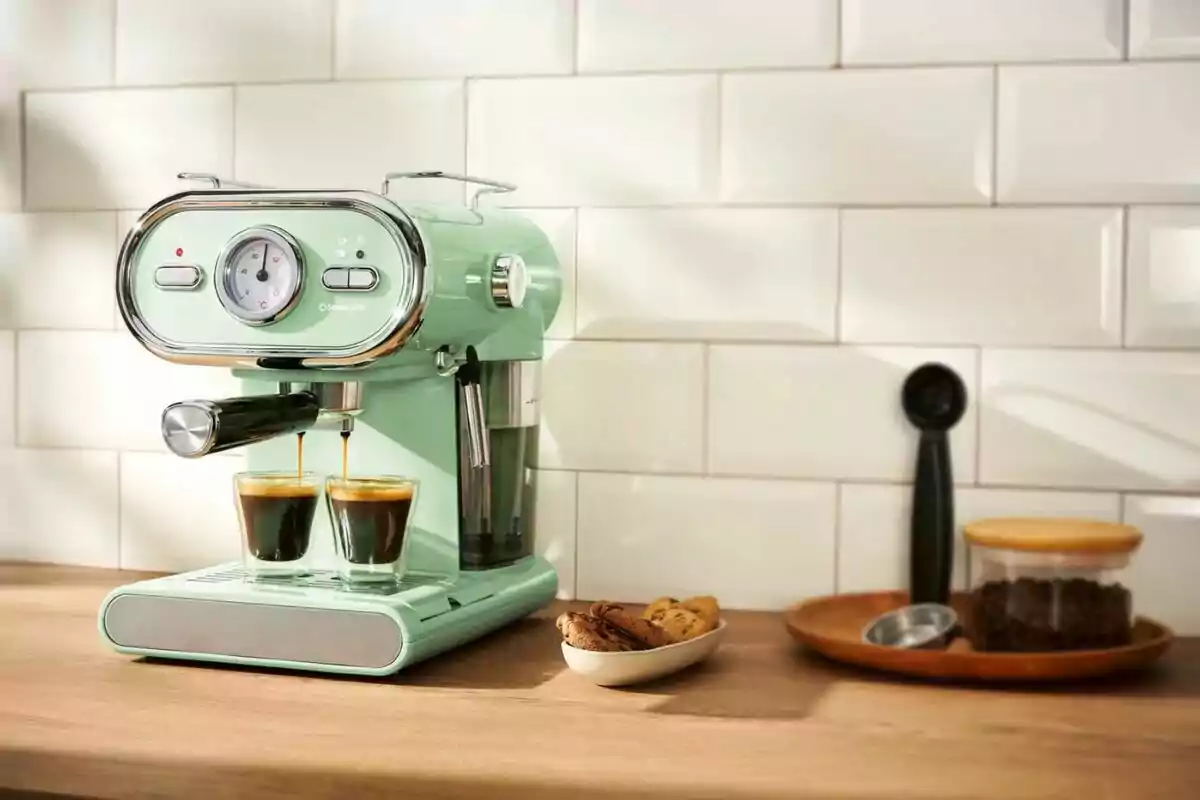 Cafetera de color verd menta preparant dos espressos en una cuina amb fons de rajoles blanques i una safata amb galetes i cafè mòlt al costat. Cafetera de color verd menta preparant dos espressos en una cuina amb fons de rajoles blanques i una safata amb galetes i cafè mòlt al costat.