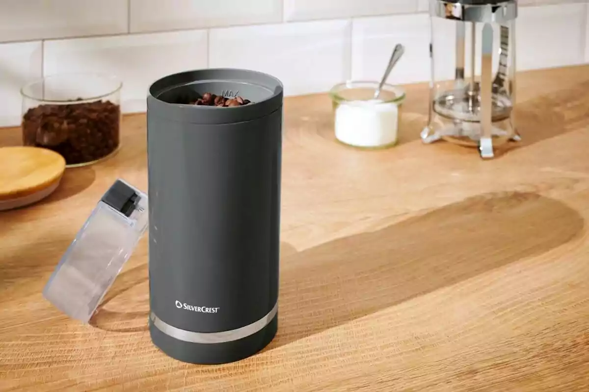 Molinillo eléctrico de café color gris sobre una encimera de madera con granos de café en su interior y una tapa transparente abierta, al fondo hay una prensa francesa, un frasco de azúcar y un recipiente con granos de café. Molinillo eléctrico de café color gris sobre una encimera de madera con granos de café en su interior y una tapa transparente abierta, al fondo hay una prensa francesa, un frasco de azúcar y un recipiente con granos de café.