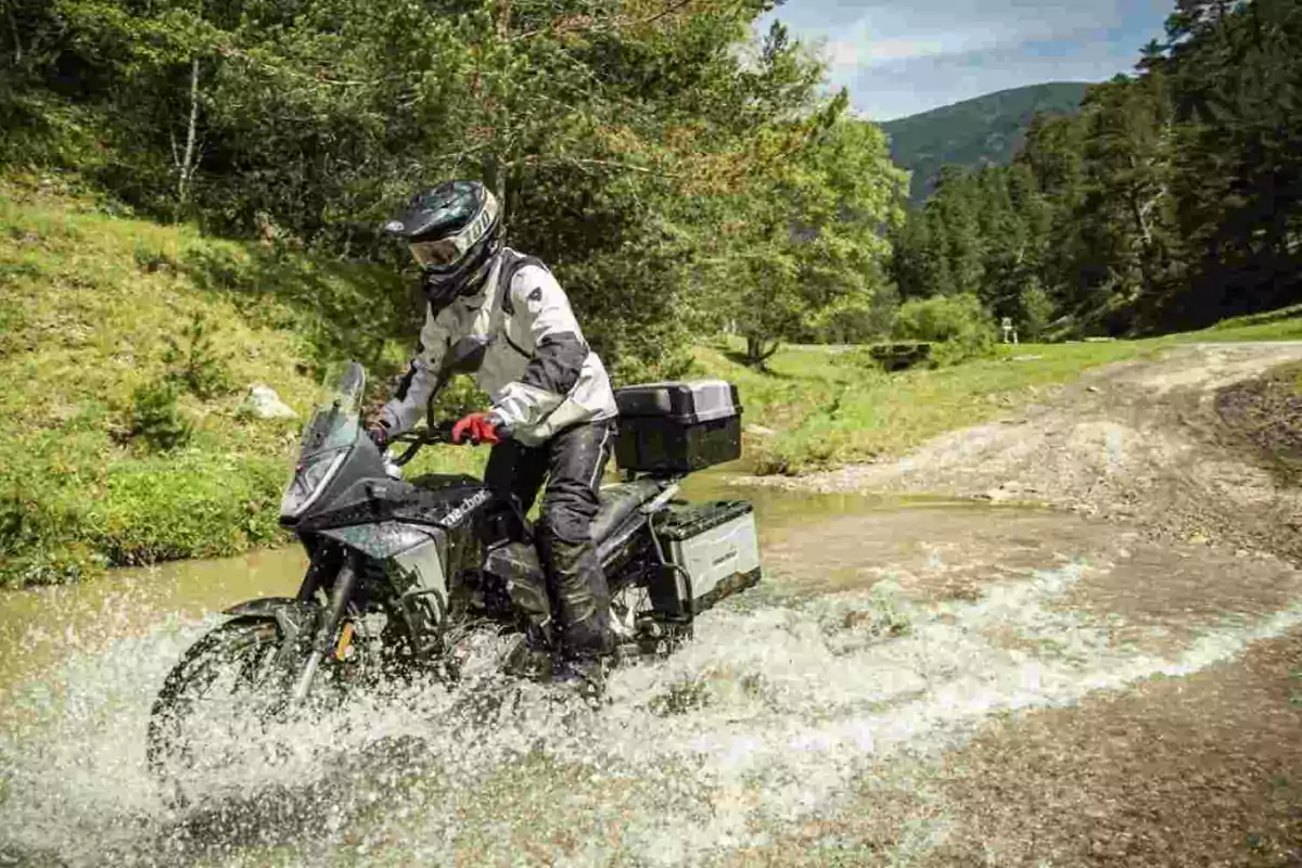 Persona amb equip de protecció conduint una motocicleta d’aventura a través d’un bassal en un entorn natural envoltat d’arbres i muntanyes