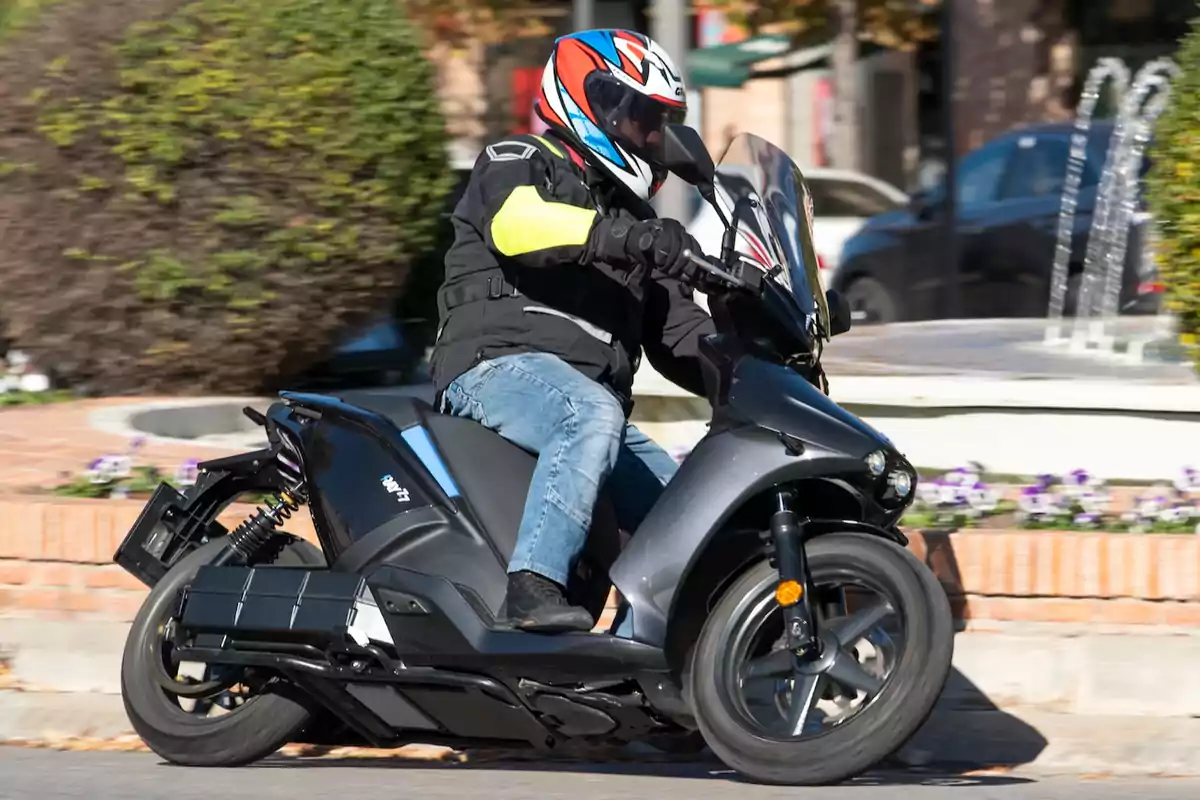 Persona conduciendo una moto eléctrica negra en una calle urbana con vegetación y una fuente al fondo Persona conduciendo una moto eléctrica negra en una calle urbana con vegetación y una fuente al fondo