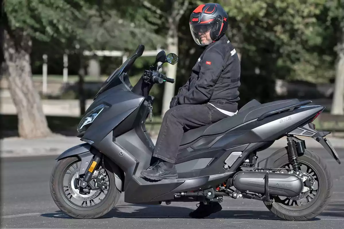 Persona con casco y chaqueta negra sentada sobre una motocicleta tipo scooter gris en una calle con árboles al fondo Persona con casco y chaqueta negra sentada sobre una motocicleta tipo scooter gris en una calle con árboles al fondo