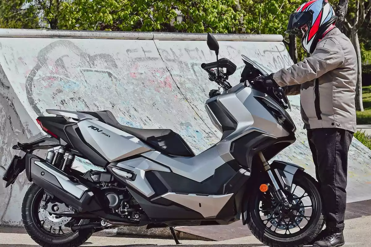 Persona con casco y chaqueta junto a una motocicleta gris estacionada frente a una rampa de skate con grafitis Persona con casco y chaqueta junto a una motocicleta gris estacionada frente a una rampa de skate con grafitis
