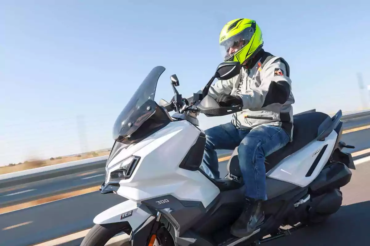 Persona conduciendo una moto tipo scooter blanca en una carretera con casco amarillo y chaqueta gris Persona conduciendo una moto tipo scooter blanca en una carretera con casco amarillo y chaqueta gris