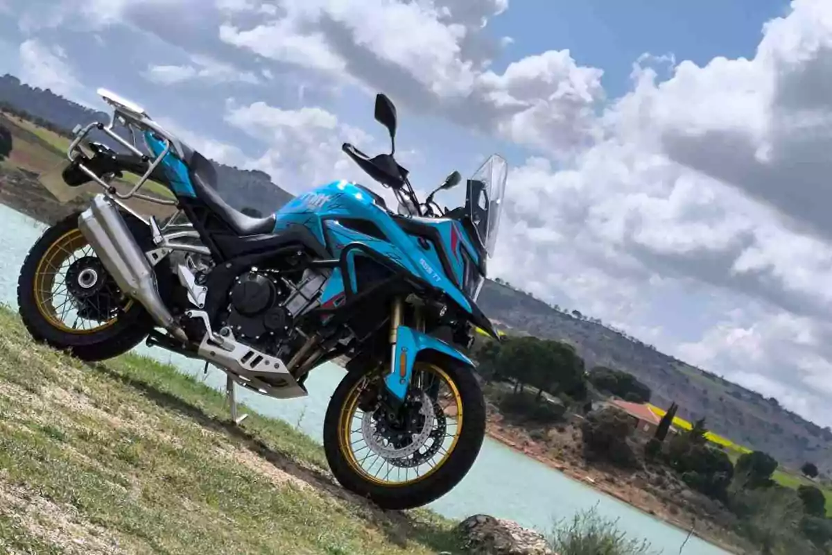 Motocicleta azul estacionada sobre césped junto a un lago con colinas y cielo nublado al fondo Motocicleta azul estacionada sobre césped junto a un lago con colinas y cielo nublado al fondo