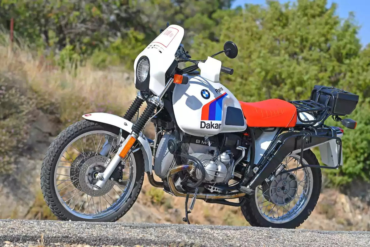 Motocicleta BMW Paris Dakar clásica con asiento rojo y detalles en azul y rojo estacionada al aire libre sobre un camino de tierra con vegetación de fondo Motocicleta BMW Paris Dakar clásica con asiento rojo y detalles en azul y rojo estacionada al aire libre sobre un camino de tierra con vegetación de fondo