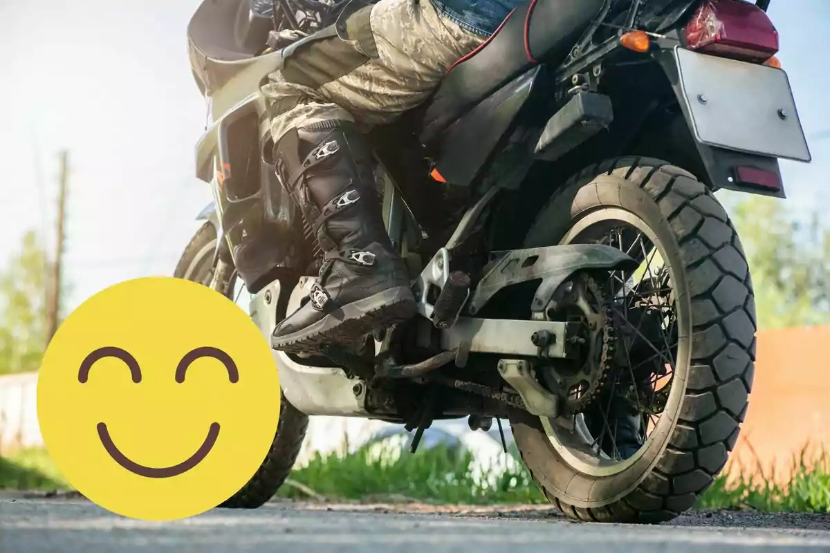 Persona con botas y pantalón camuflado sentada en una motocicleta negra junto a un emoji amarillo sonriente