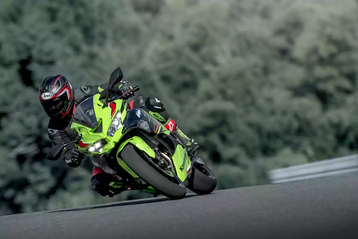 Motociclista con casco y traje de protección conduciendo una motocicleta deportiva verde en una curva de pista asfaltada con fondo de árboles desenfocados Motociclista con casco y traje de protección conduciendo una motocicleta deportiva verde en una curva de pista asfaltada con fondo de árboles desenfocados
