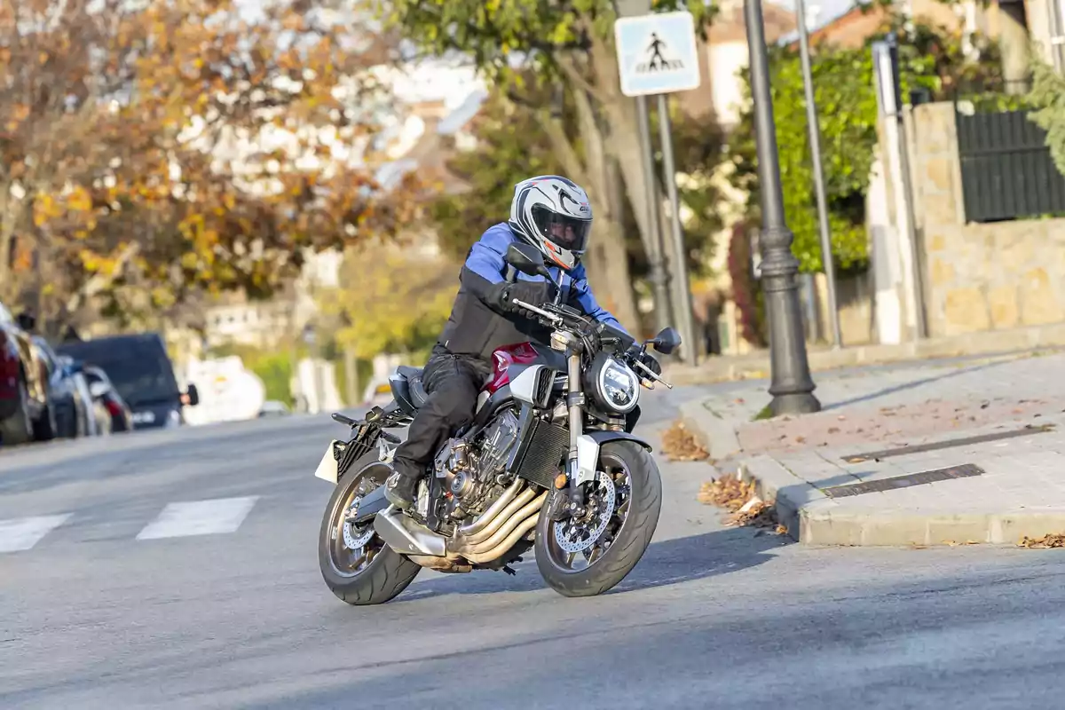 Persona con casco y chaqueta azul conduciendo una motocicleta en una calle urbana con árboles y señal de cruce peatonal al fondo Persona con casco y chaqueta azul conduciendo una motocicleta en una calle urbana con árboles y señal de cruce peatonal al fondo