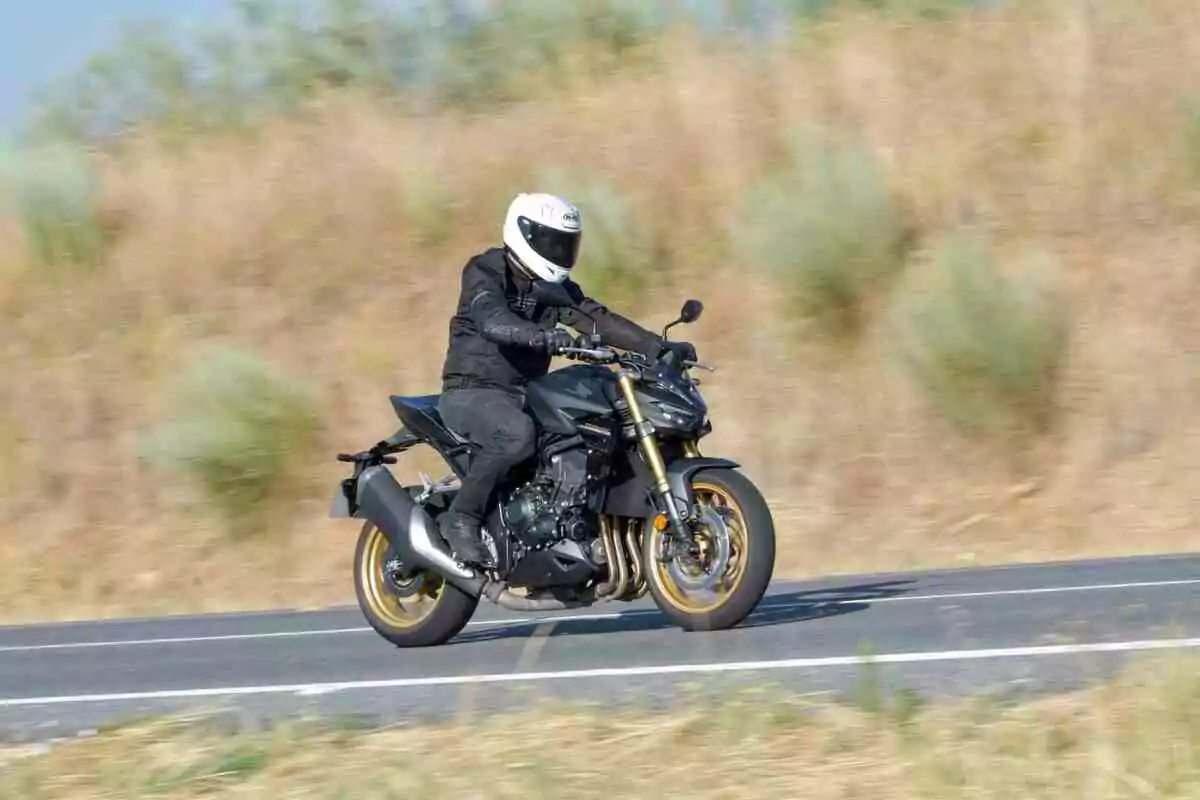 Persona con casco blanco y ropa negra conduciendo una motocicleta negra en una carretera asfaltada con vegetación seca al fondo Persona con casco blanco y ropa negra conduciendo una motocicleta negra en una carretera asfaltada con vegetación seca al fondo