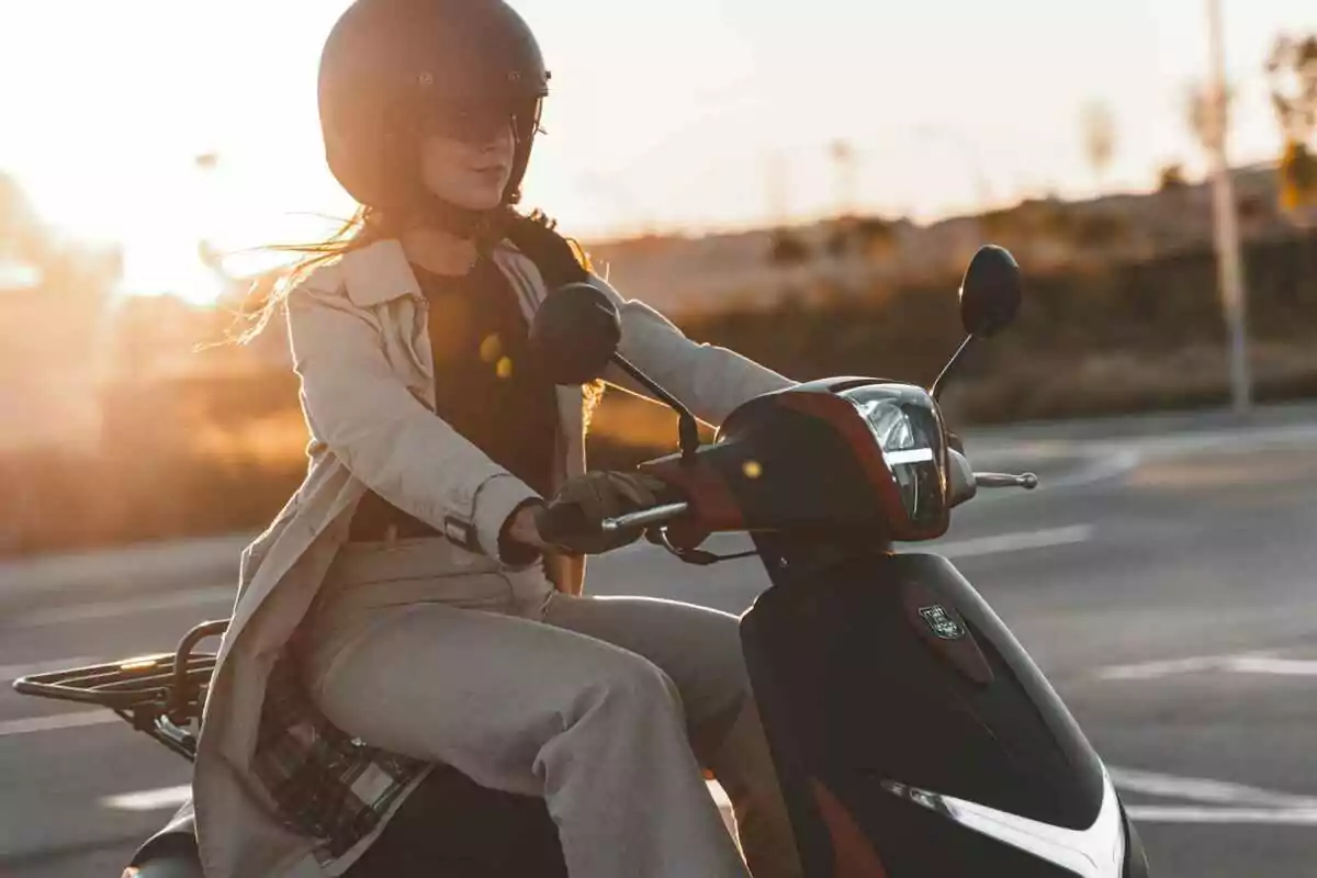 Mujer conduciendo una motoneta negra al atardecer usando casco y gafas de sol Mujer conduciendo una motoneta negra al atardecer usando casco y gafas de sol