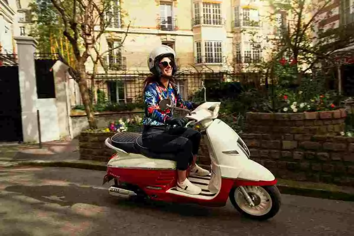 Mujer sonriente conduciendo una motoneta blanca y roja por una calle urbana con flores y edificios al fondo Mujer sonriente conduciendo una motoneta blanca y roja por una calle urbana con flores y edificios al fondo