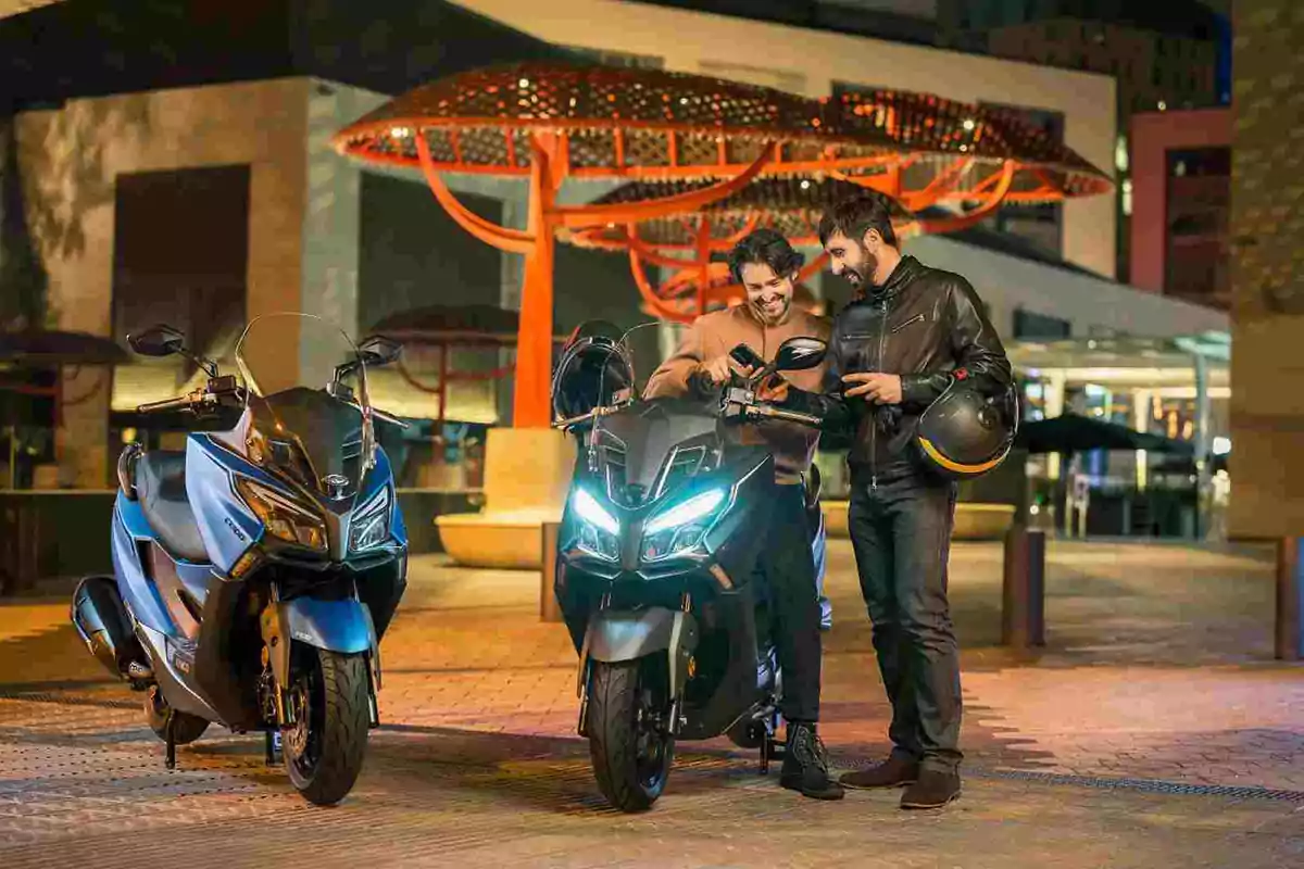 Dos hombres sonríen junto a dos motocicletas encendidas en una ciudad iluminada por la noche Dos hombres sonríen junto a dos motocicletas encendidas en una ciudad iluminada por la noche
