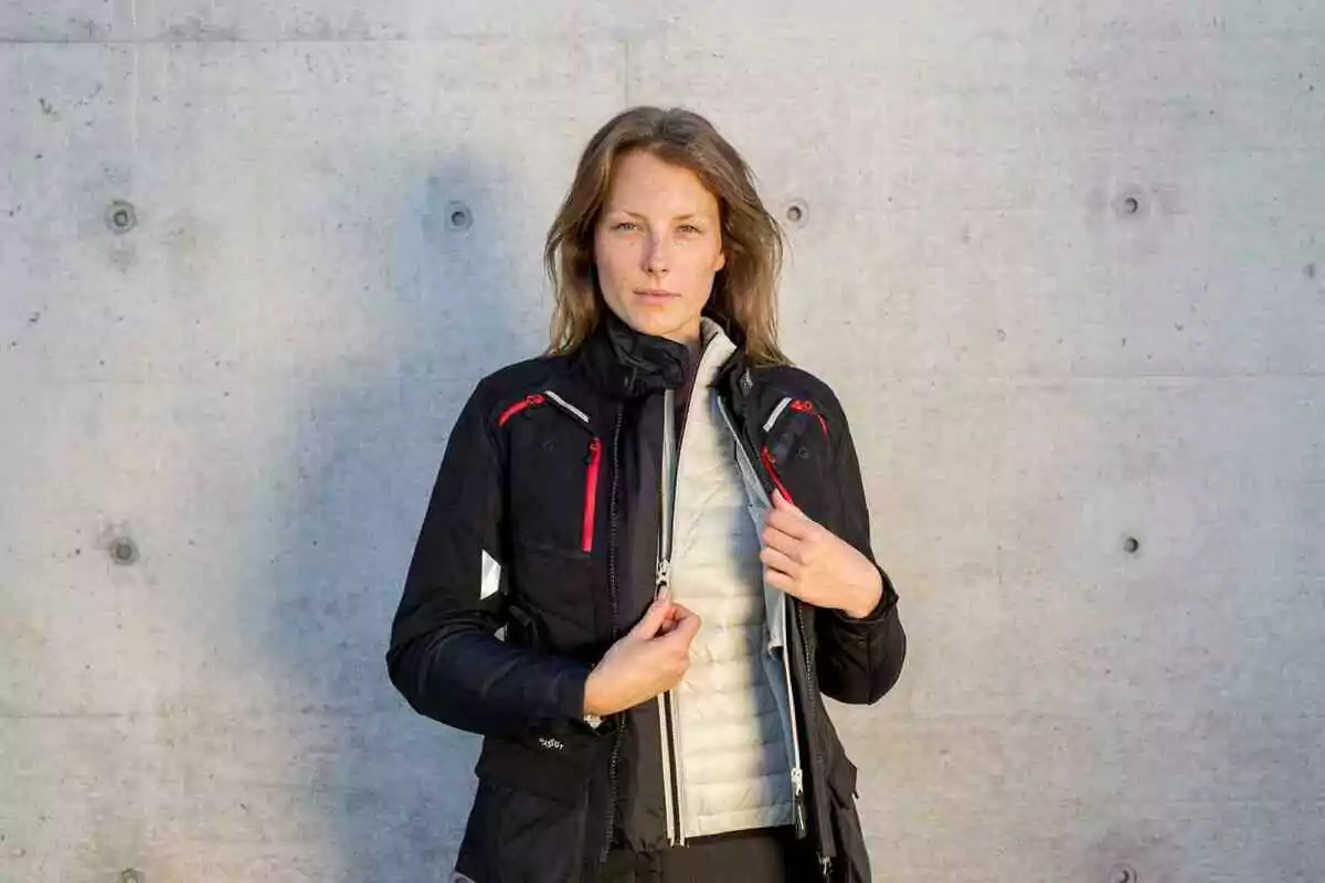 Mujer de cabello castaño claro usando una chaqueta negra con detalles rojos y un chaleco claro debajo, de pie frente a una pared de concreto Mujer de cabello castaño claro usando una chaqueta negra con detalles rojos y un chaleco claro debajo, de pie frente a una pared de concreto