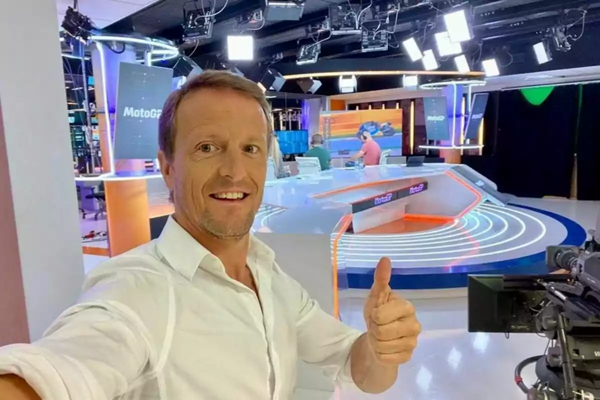 Hombre sonriente con camisa blanca haciendo un gesto de pulgar arriba en un moderno estudio de televisión con pantallas y luces, donde se ve el logo de MotoGP en el fondo Hombre sonriente con camisa blanca haciendo un gesto de pulgar arriba en un moderno estudio de televisión con pantallas y luces, donde se ve el logo de MotoGP en el fondo