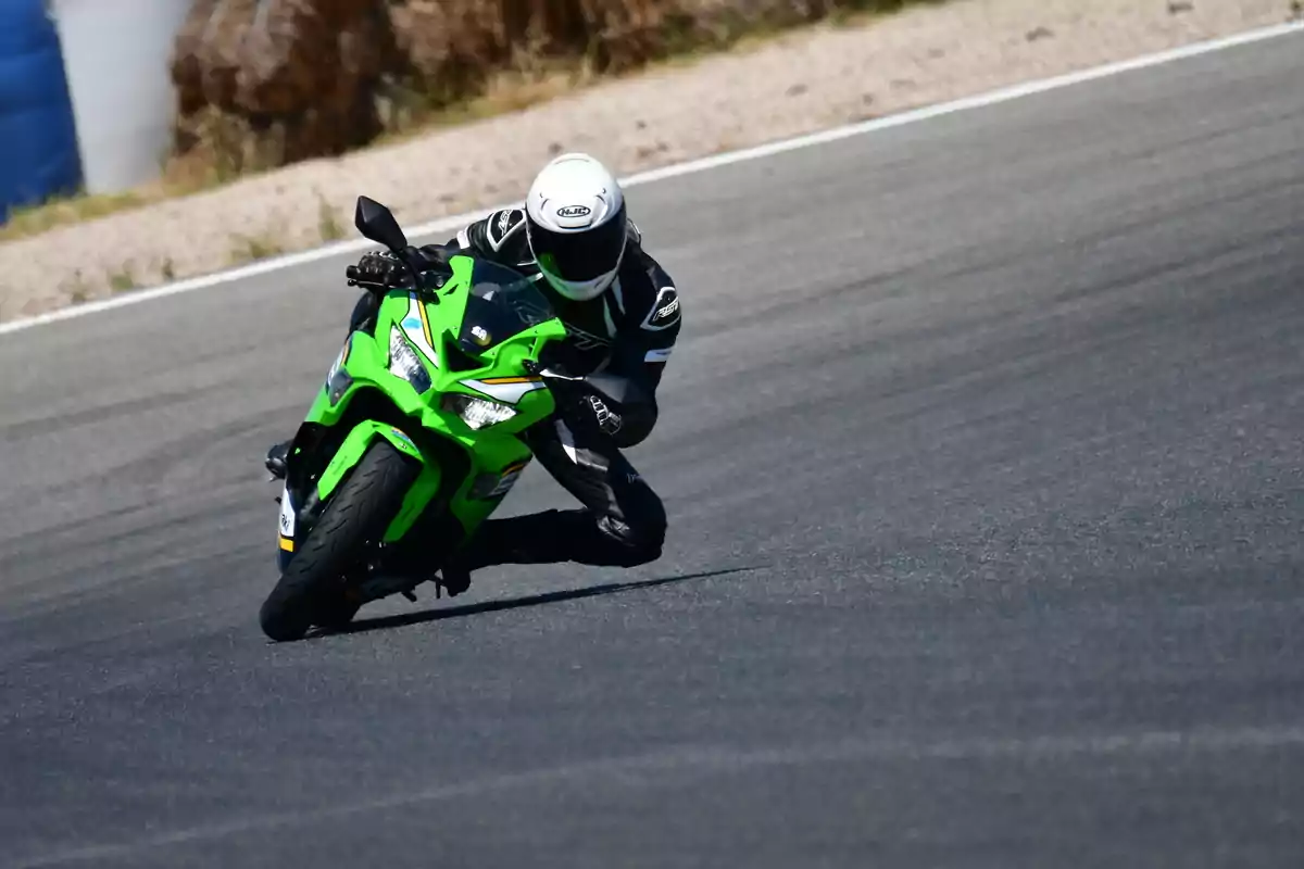 Motociclista con casco blanco y traje negro tomando una curva cerrada en una pista de carreras sobre una motocicleta deportiva verde Motociclista con casco blanco y traje negro tomando una curva cerrada en una pista de carreras sobre una motocicleta deportiva verde