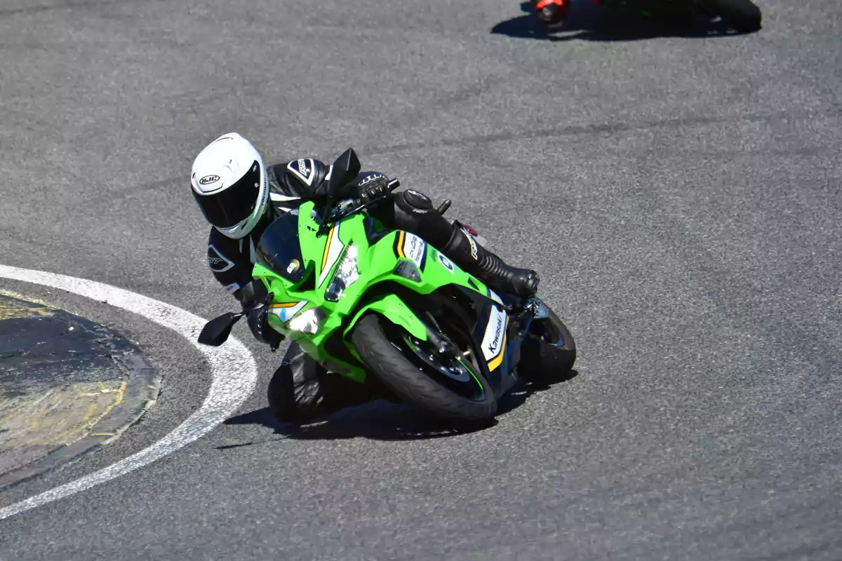 Motociclista con casco blanco y traje negro tomando una curva cerrada en una motocicleta deportiva verde sobre una pista de carreras asfaltada Motociclista con casco blanco y traje negro tomando una curva cerrada en una motocicleta deportiva verde sobre una pista de carreras asfaltada