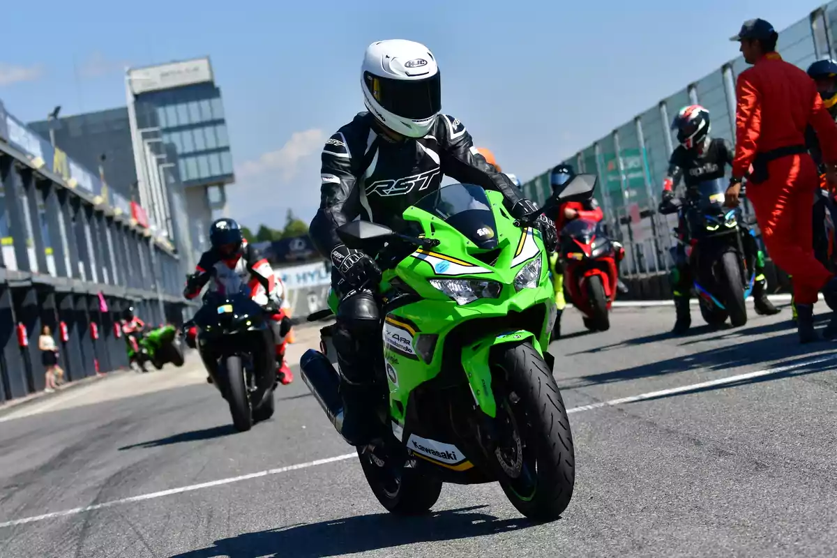 Pilotos de motocicletas deportivas en un circuito de carreras con un motociclista en una moto verde al frente y varios más al fondo junto a personal de pista Pilotos de motocicletas deportivas en un circuito de carreras con un motociclista en una moto verde al frente y varios más al fondo junto a personal de pista