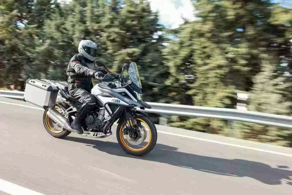 Persona conduciendo una motocicleta de turismo equipada con maletas laterales en una carretera rodeada de árboles