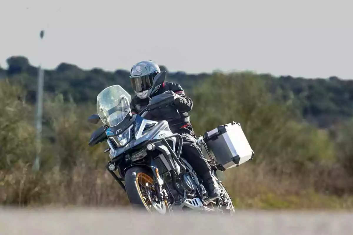 Persona con casco y equipo de protección conduciendo una motocicleta de aventura en una carretera rodeada de vegetación. Persona con casco y equipo de protección conduciendo una motocicleta de aventura en una carretera rodeada de vegetación.