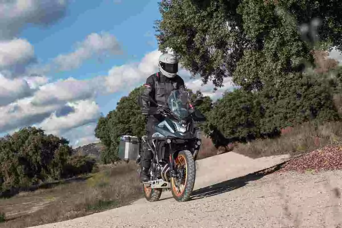 Persona con casco y equipo de protección conduciendo una motocicleta por un camino de tierra rodeado de árboles y cielo parcialmente nublado Persona con casco y equipo de protección conduciendo una motocicleta por un camino de tierra rodeado de árboles y cielo parcialmente nublado
